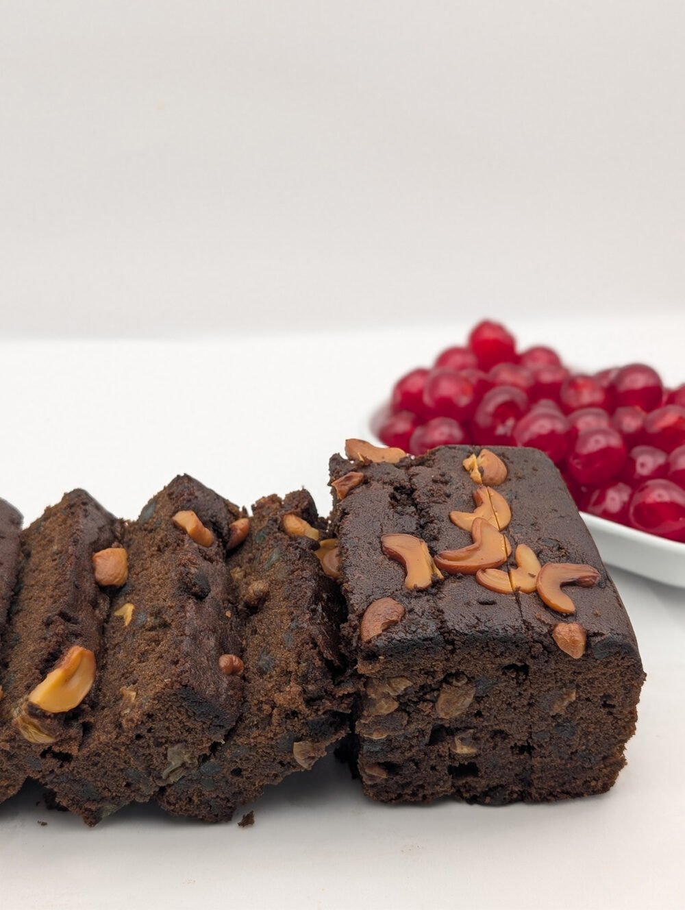 Slices of rich plum cake topped with golden cashews, with glazed red cherries in the background
