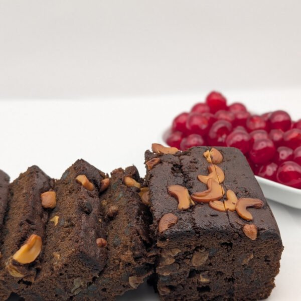 Slices of rich plum cake topped with golden cashews, with glazed red cherries in the background