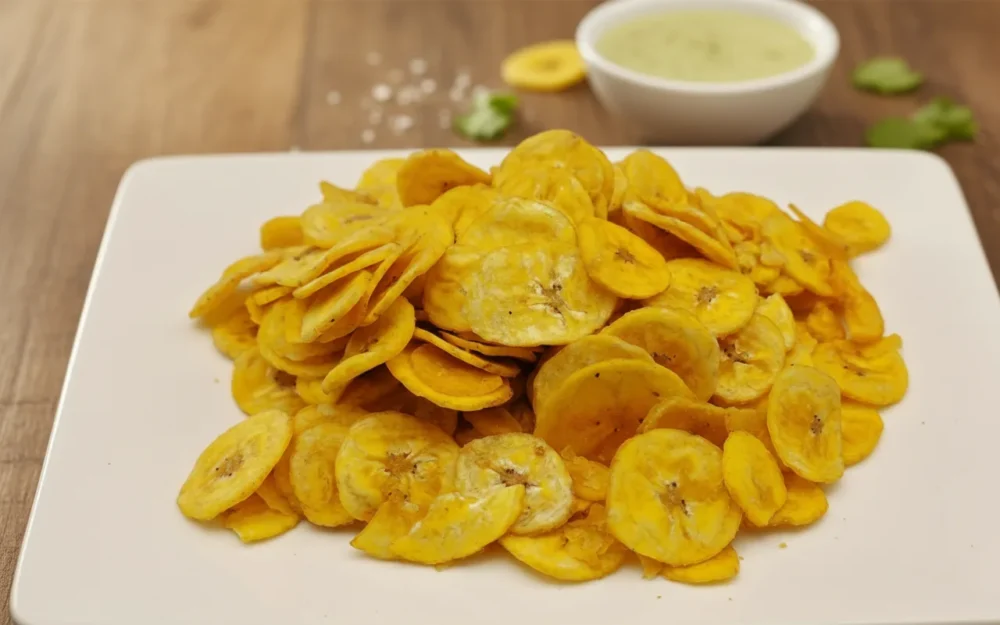 Golden fried Mangalorean banana chips piled high on a square white plate, set on a wooden table with a side of green chutney and coarse salt, traditional Christmas Kuswar savoury snack.