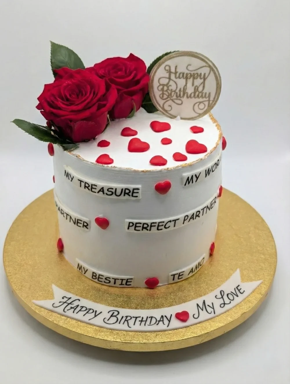 Small, round white cake decorated with red roses, red heart cutouts, a gold 'Happy Birthday' topper, and multiple small banners with phrases like "MY TREASURE," "PERFECT PARTNER," and "TE AMO." A larger banner on the gold cake board reads "HAPPY BIRTHDAY MY LOVE."