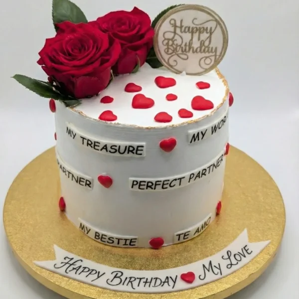 Small, round white cake decorated with red roses, red heart cutouts, a gold 'Happy Birthday' topper, and multiple small banners with phrases like "MY TREASURE," "PERFECT PARTNER," and "TE AMO." A larger banner on the gold cake board reads "HAPPY BIRTHDAY MY LOVE."