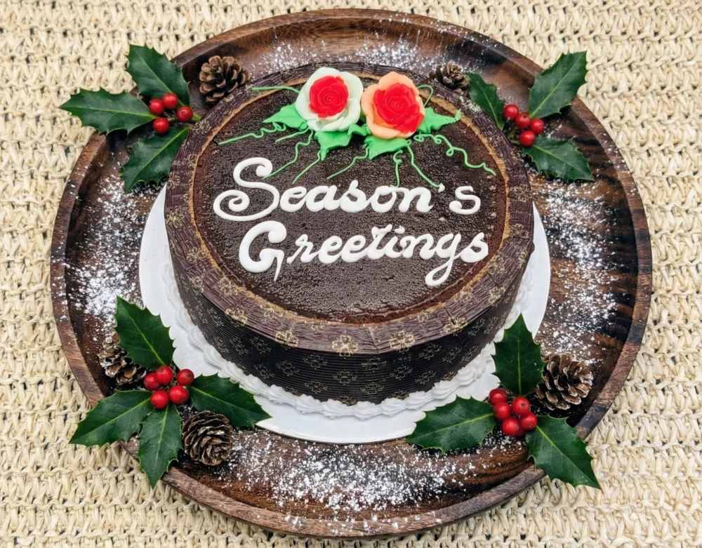 Top-down view of a round dark Christmas plum cake on a wooden platter. The cake features white icing text reading 'Season's Greetings' and is decorated with red and peach fondant roses and green vines. The platter is garnished with holly leaves, pinecones, and powdered sugar snow.