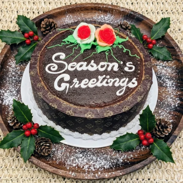 Top-down view of a round dark Christmas plum cake on a wooden platter. The cake features white icing text reading 'Season's Greetings' and is decorated with red and peach fondant roses and green vines. The platter is garnished with holly leaves, pinecones, and powdered sugar snow.