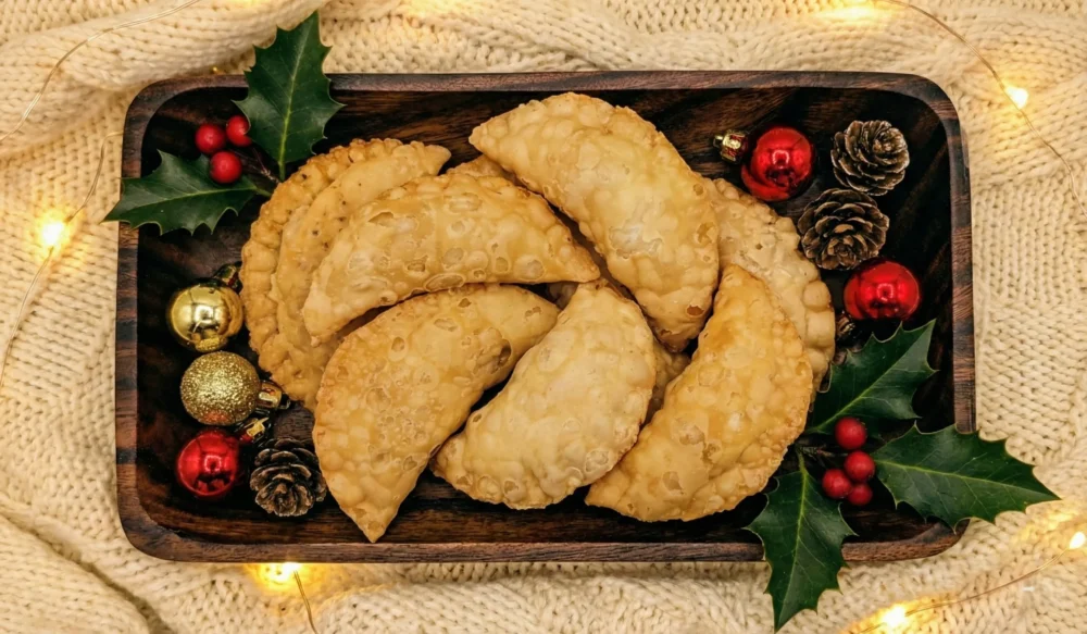 Overhead view of a wooden rectangular platter filled with golden-fried Mangalorean Newri pastries (sweet crescent dumplings). The platter is decorated with festive green holly leaves, red berries, small pinecones, and shiny red and gold Christmas ornaments, set against a cozy cream knitted background.