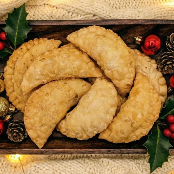 Overhead view of a wooden rectangular platter filled with golden-fried Mangalorean Newri pastries (sweet crescent dumplings). The platter is decorated with festive green holly leaves, red berries, small pinecones, and shiny red and gold Christmas ornaments, set against a cozy cream knitted background.