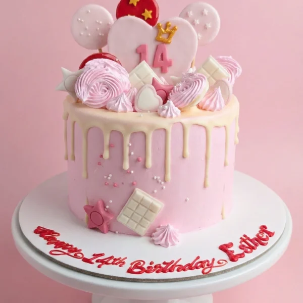 Pastel pink drip birthday cake for 14th birthday featuring white chocolate drip, pink meringue swirls, chocolate squares, and a heart-shaped topper with a gold crown. Cake board reads Happy 14th Birthday Esther in red script.