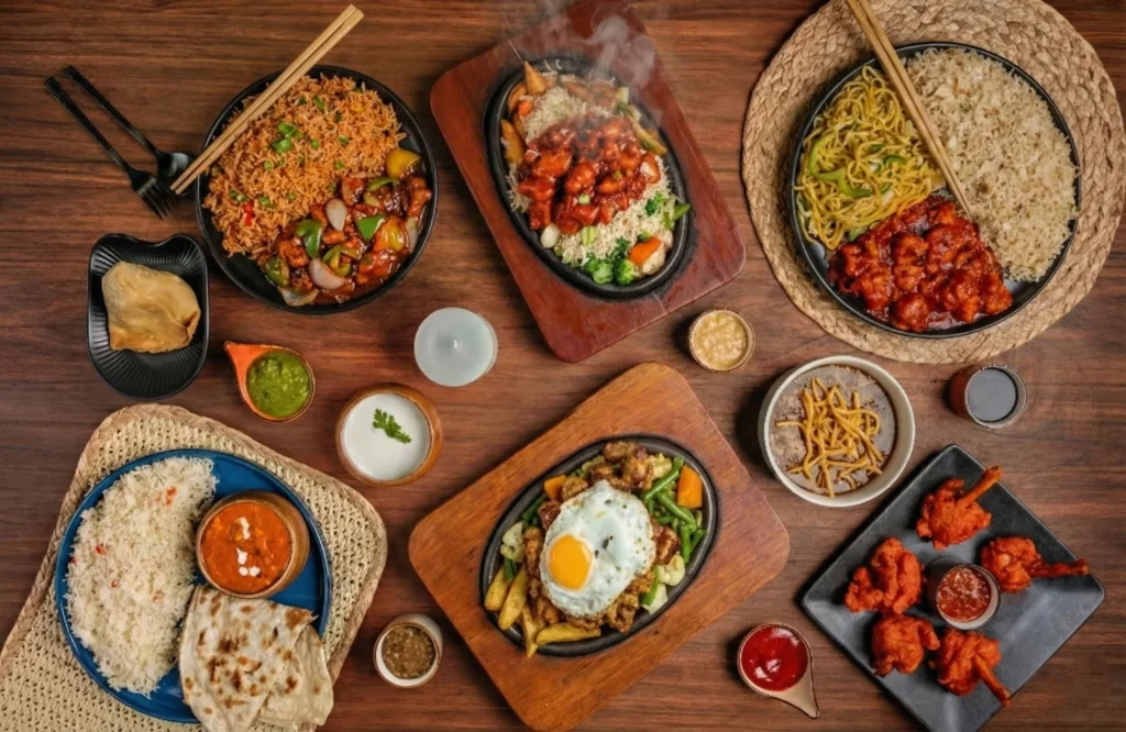 Overhead view of assorted Indo-Chinese and Indian dishes including chicken sizzlers, fried rice, noodles, curries, naan, and crispy appetizers served on wooden platters and bowls.