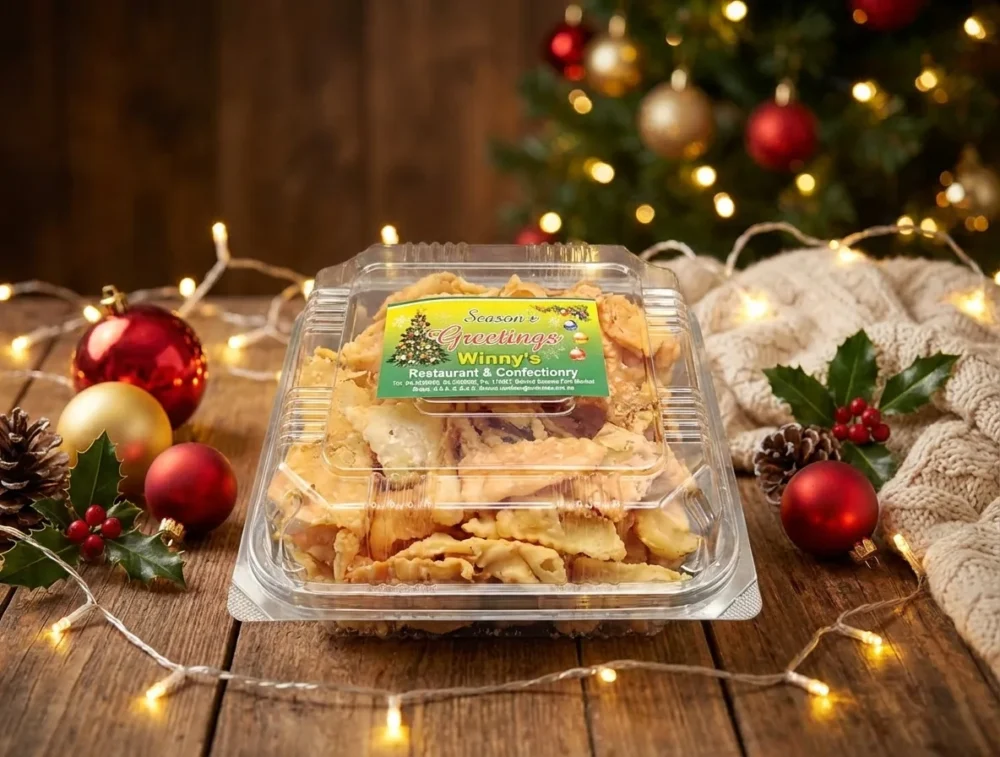 High-angle view of a clear plastic container packed with crispy golden Thukudi snacks, featuring a 'Season's Greetings' label from Winny's. The box is arranged on a rustic wooden table surrounded by festive holiday decor, including pinecones, red holly berries, and warm glowing Christmas lights.