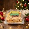 High-angle view of a clear plastic container packed with crispy golden Thukudi snacks, featuring a 'Season's Greetings' label from Winny's. The box is arranged on a rustic wooden table surrounded by festive holiday decor, including pinecones, red holly berries, and warm glowing Christmas lights.