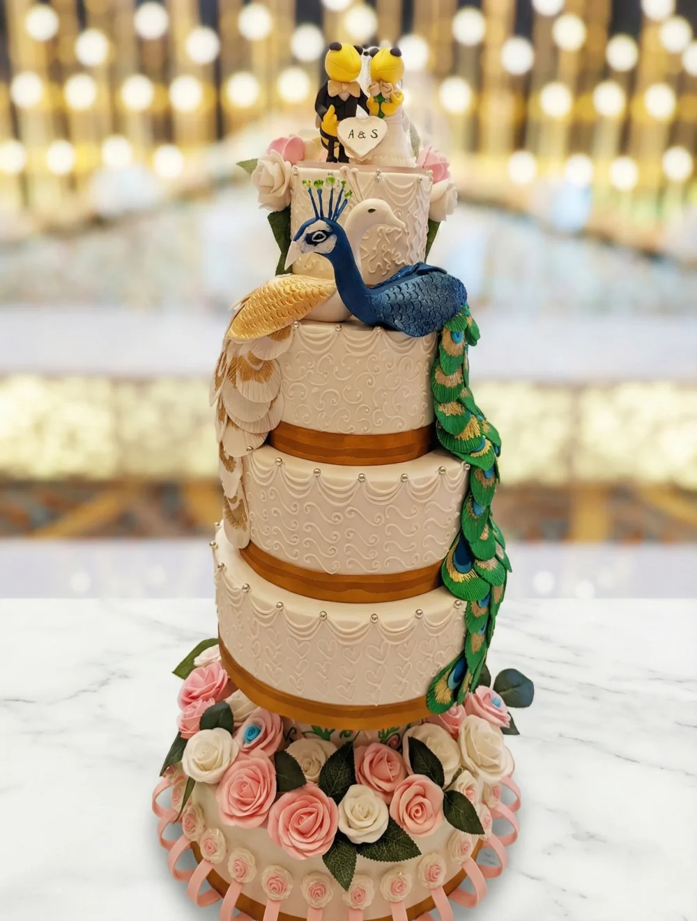 A four-tier white wedding cake with gold accents, decorated with a blue and green peacock and a gold peacock whose tails cascade down the side, sitting on a bed of pink and white roses. At the very top are two Georgia Tech yellow jacket mascots dressed as a bride and groom, holding a heart that reads "A&S."