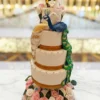 A four-tier white wedding cake with gold accents, decorated with a blue and green peacock and a gold peacock whose tails cascade down the side, sitting on a bed of pink and white roses. At the very top are two Georgia Tech yellow jacket mascots dressed as a bride and groom, holding a heart that reads "A&S."