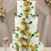 Three-tier white wedding cake featuring cascading gold and white roses, green foliage, and scattered gold leaf accents.